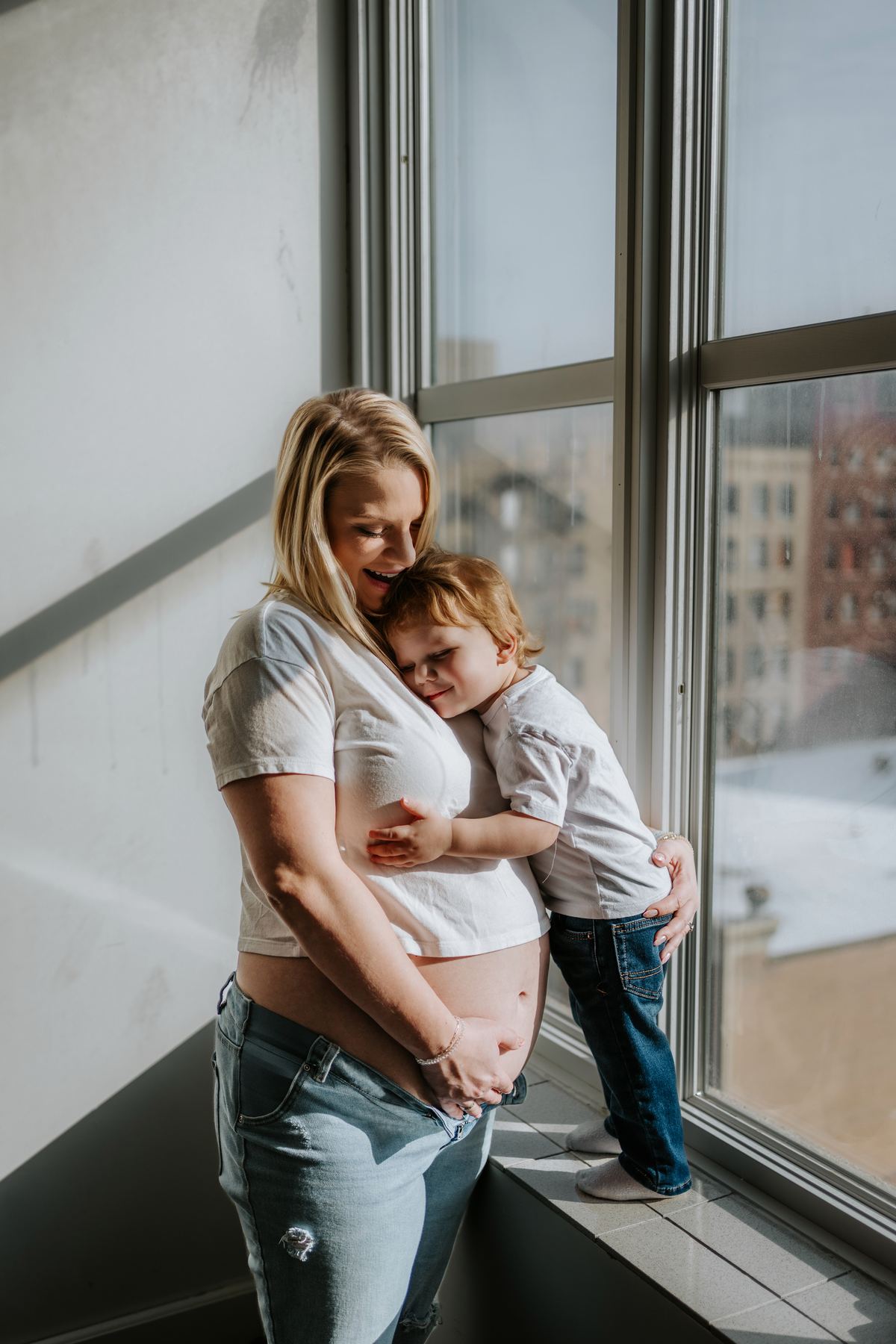 Mother and toddler embracing by a large window — warm natural light family and maternity photography in Winnipeg by Ngo Photography
