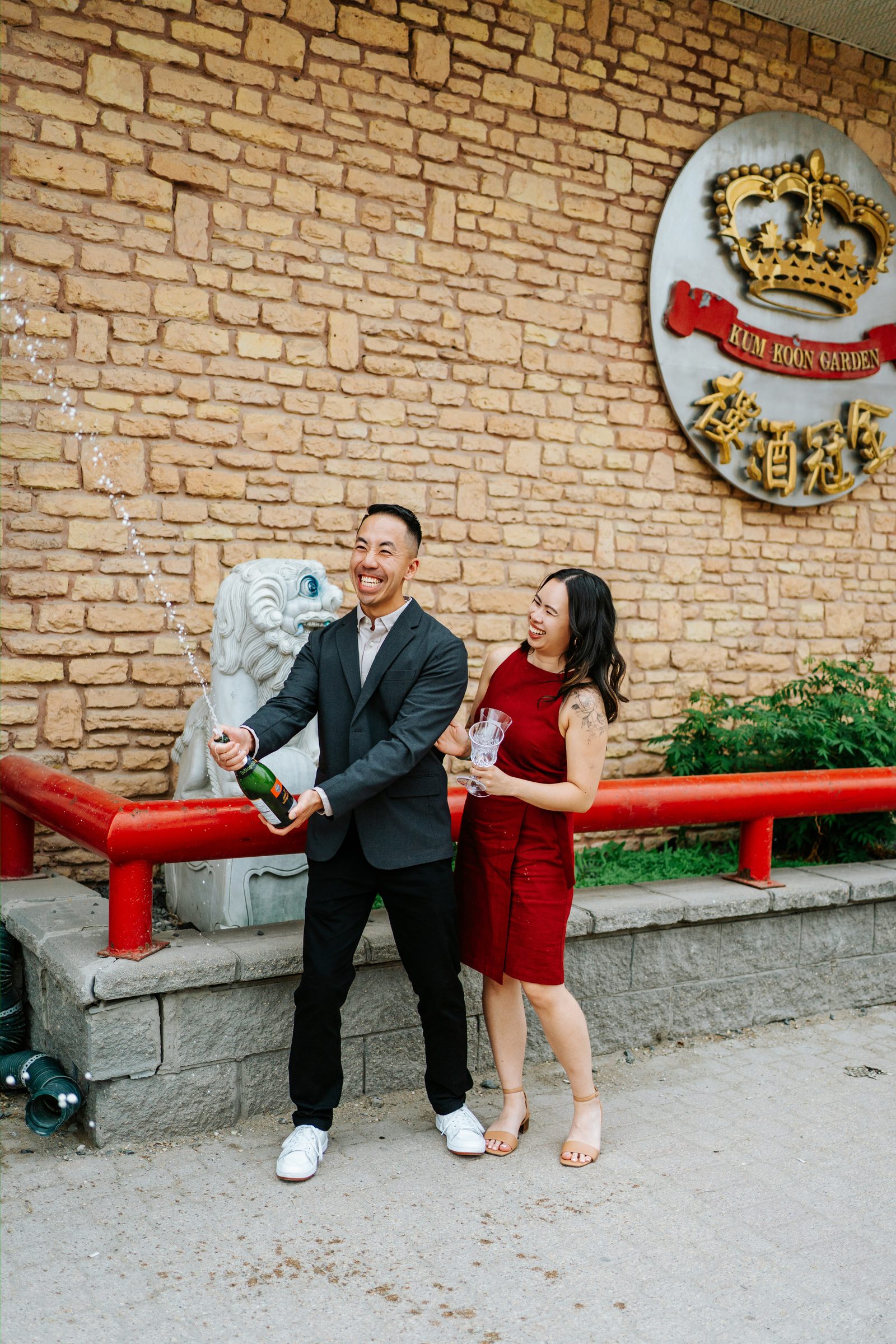 Jason popping champagne outside Kum Koon Garden Winnipeg with Monica laughing — Chinese engagement celebration photography by Ngo Photography