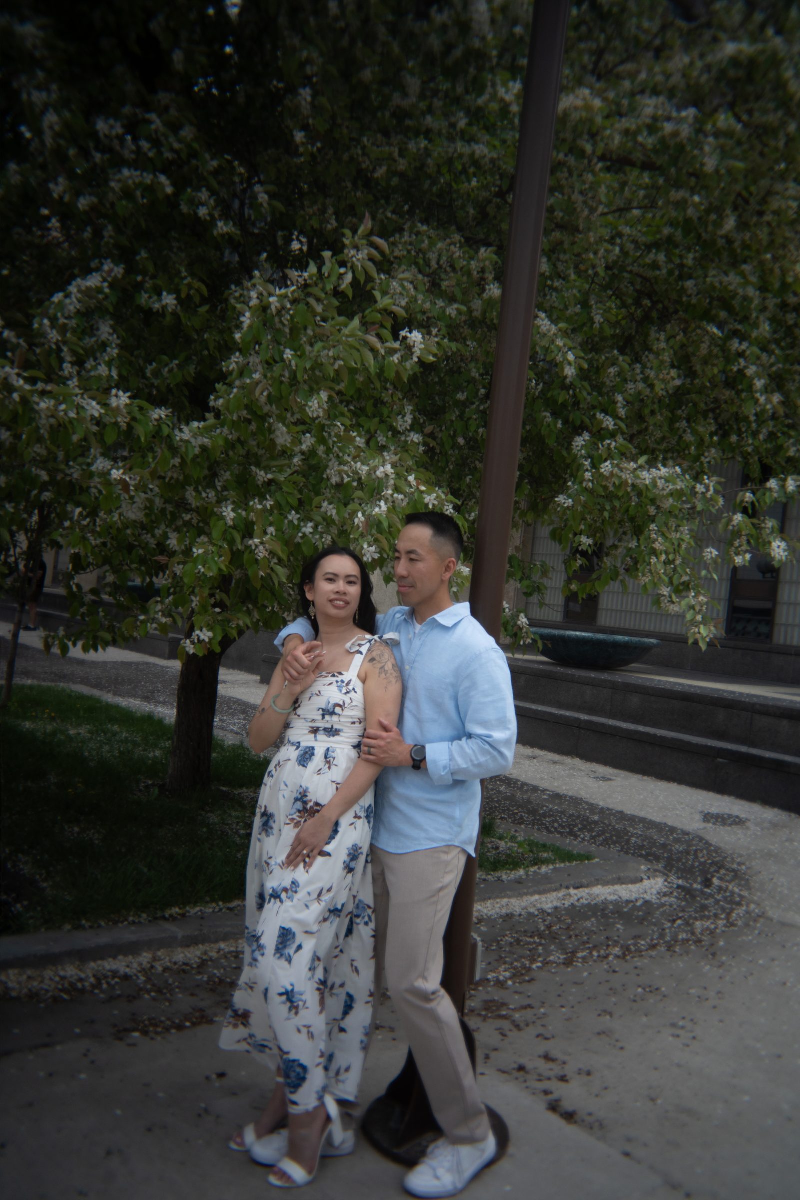 Monica and Jason embracing under blossoming trees in Winnipeg's Chinatown neighbourhood — engagement photography by Chris Ngo