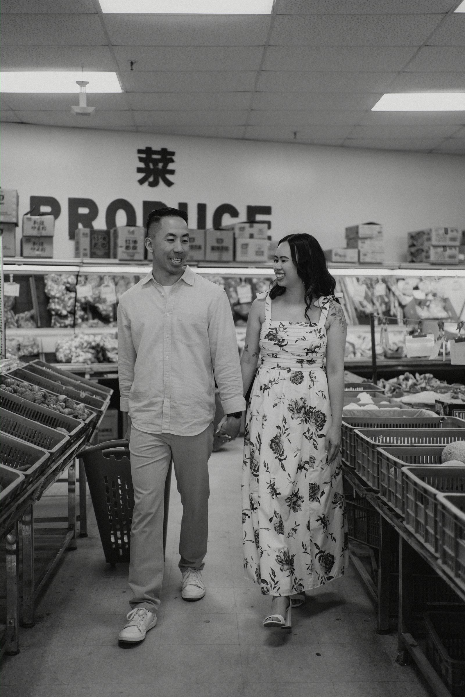 Monica and Jason walking through the produce section of Sun-Wah Centre Winnipeg with Chinese character 菜 and PRODUCE sign on the wall — black and white engagement photography by Chris Ngo