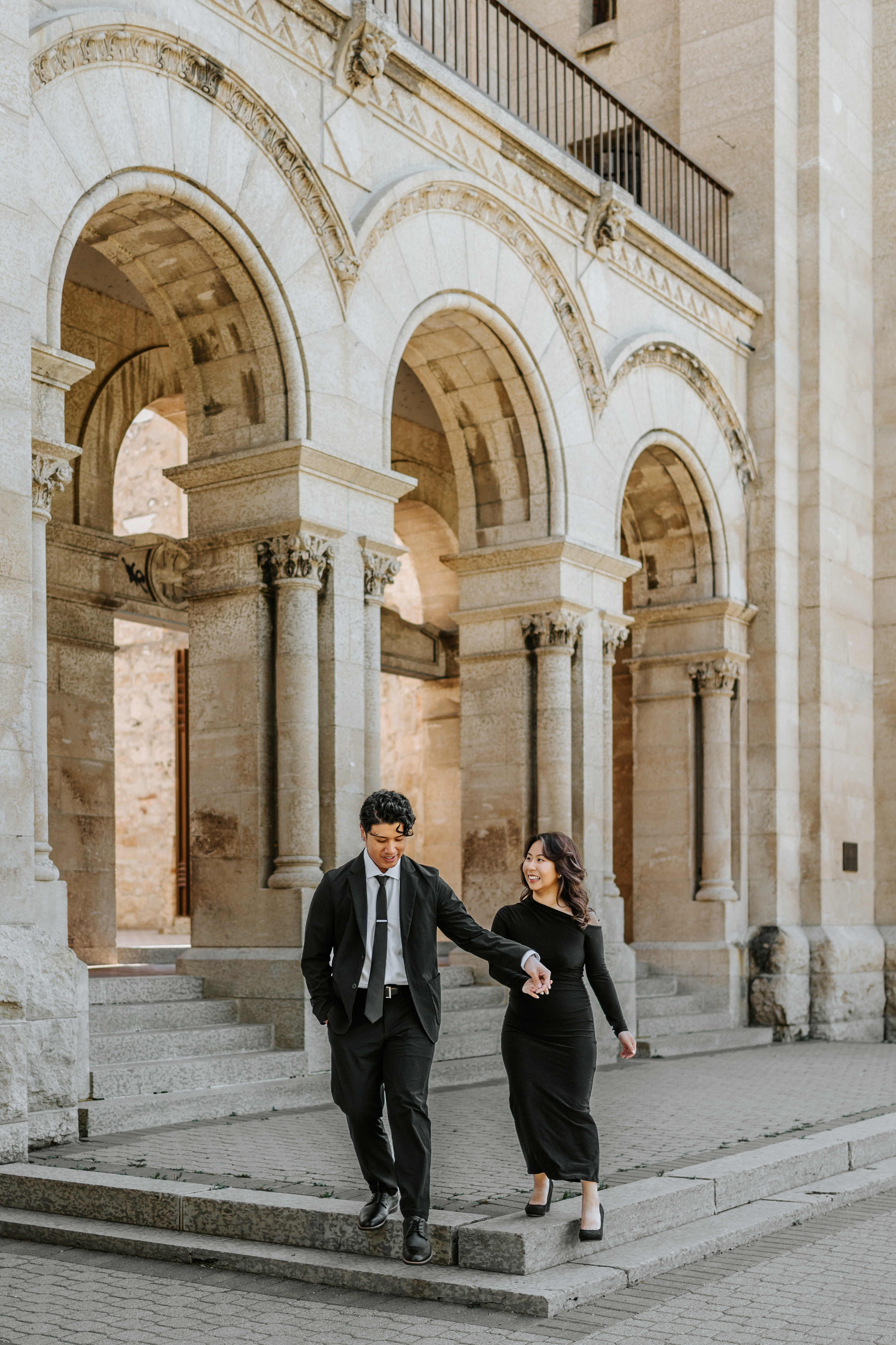 Noah and Melissa walking hand-in-hand along the stone arched colonnade at the Manitoba Legislative Building exterior — engagement photography Ngo Photography Winnipeg