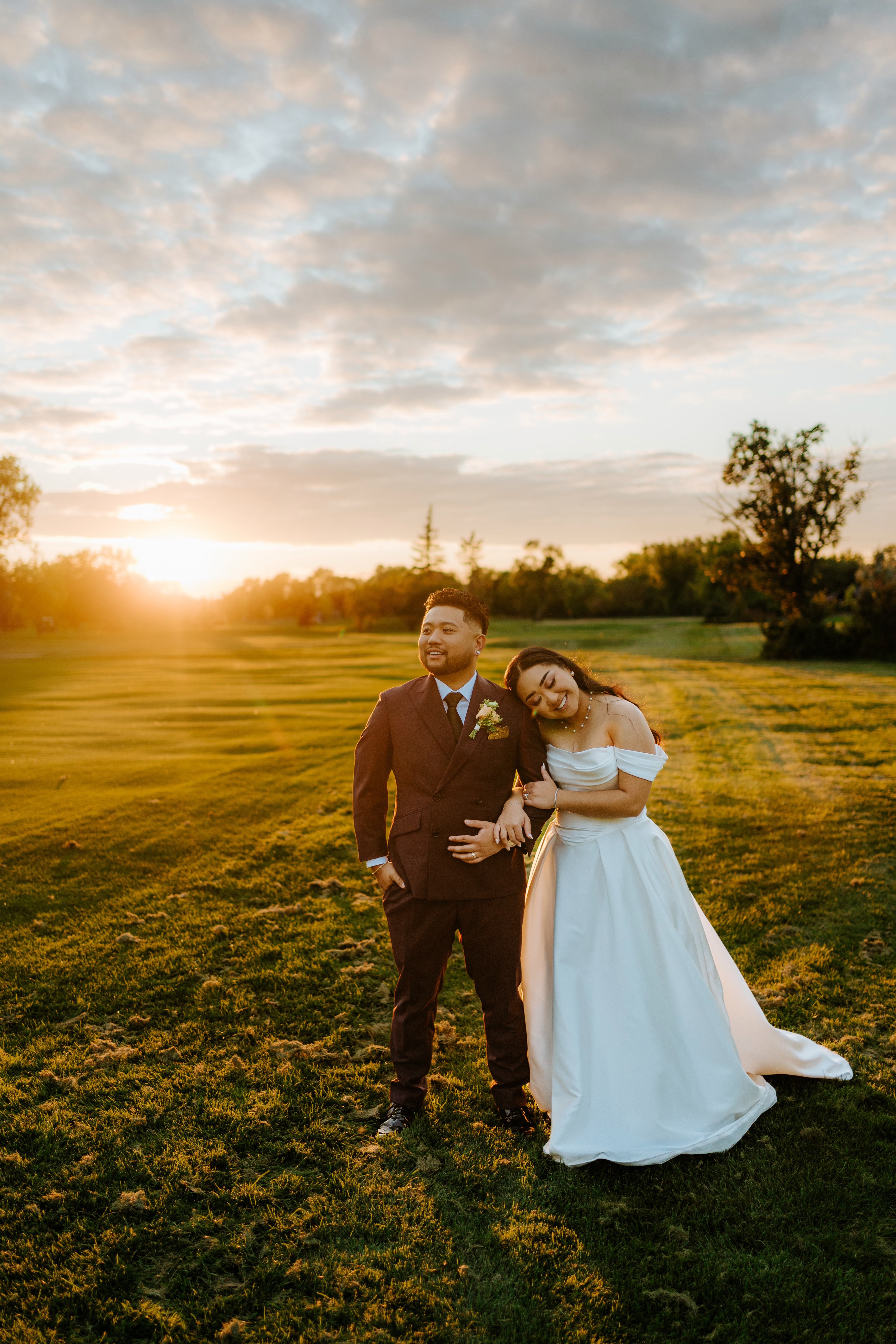 Paul and Ayanna standing together on the Breezy Bend fairway at golden hour, Ayanna leaning into Paul with a dramatic sunset and clouds behind them — Ngo Photography Winnipeg