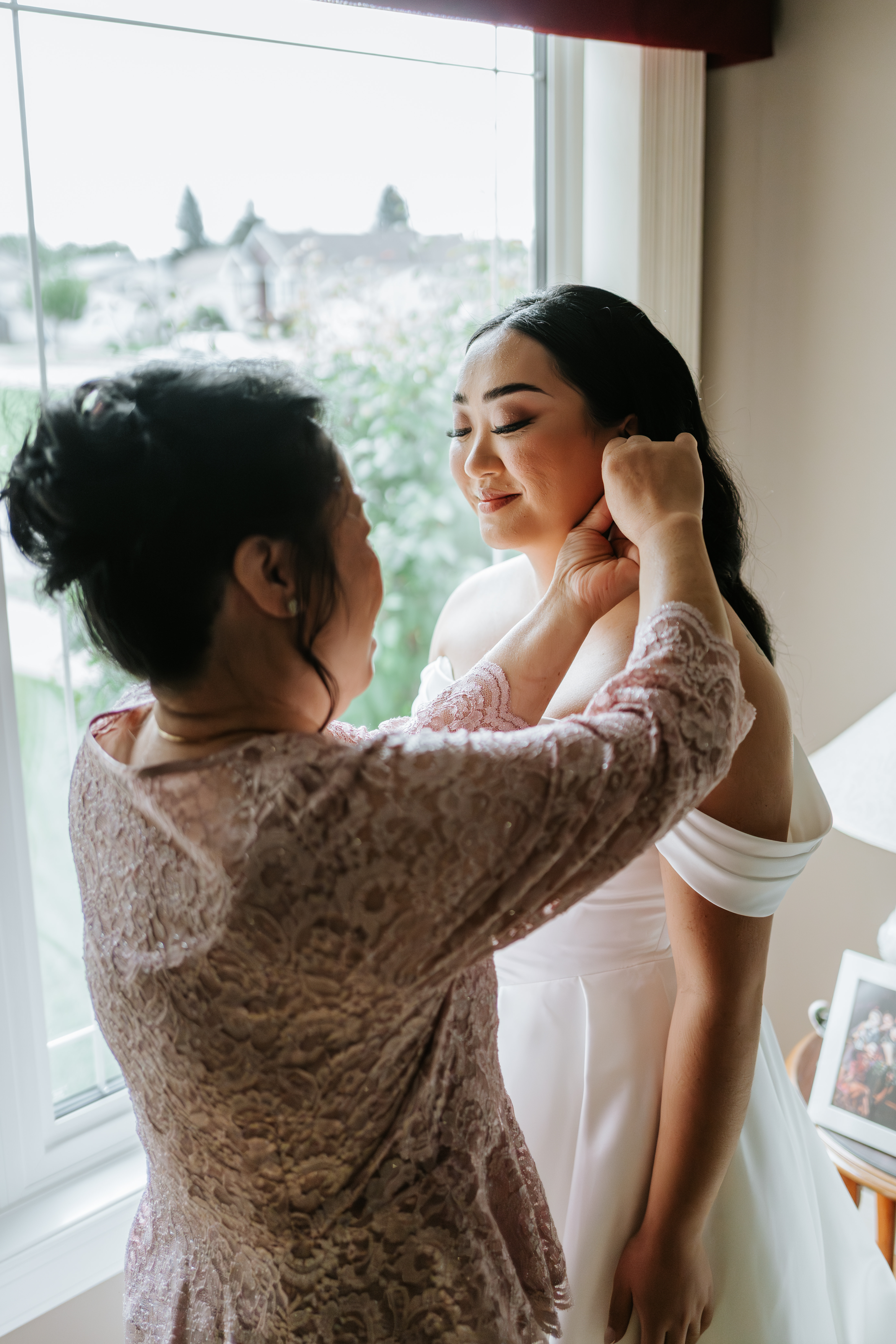 Mother adjusting Ayanna's earring as she smiles softly by the window on her wedding morning — getting ready photography by Ngo Photography Winnipeg