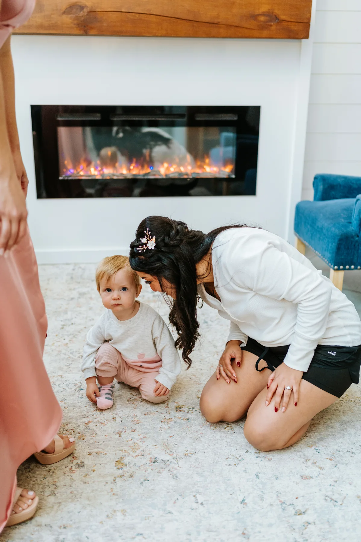 Bride Paola kneeling with a baby by the stone fireplace during getting ready at Oakland Estates Manitoba wedding