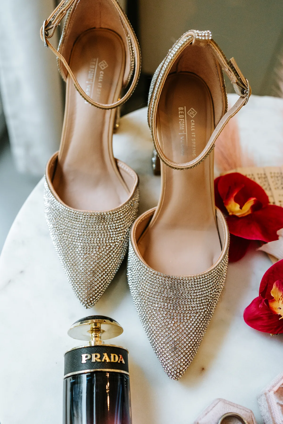 Bridal detail flat lay at Oakland Estates — champagne crystal heeled shoes, Prada perfume and red orchid blooms on marble
