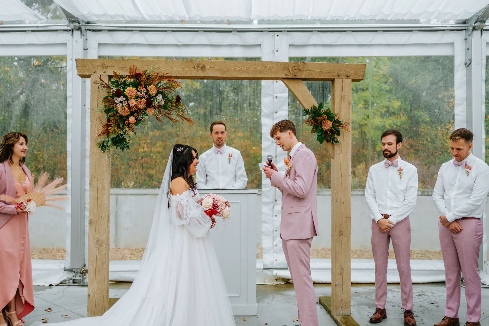Paola and Riley exchanging vows at the wooden arch inside the glass tent ceremony at Oakland Estates Manitoba wedding
