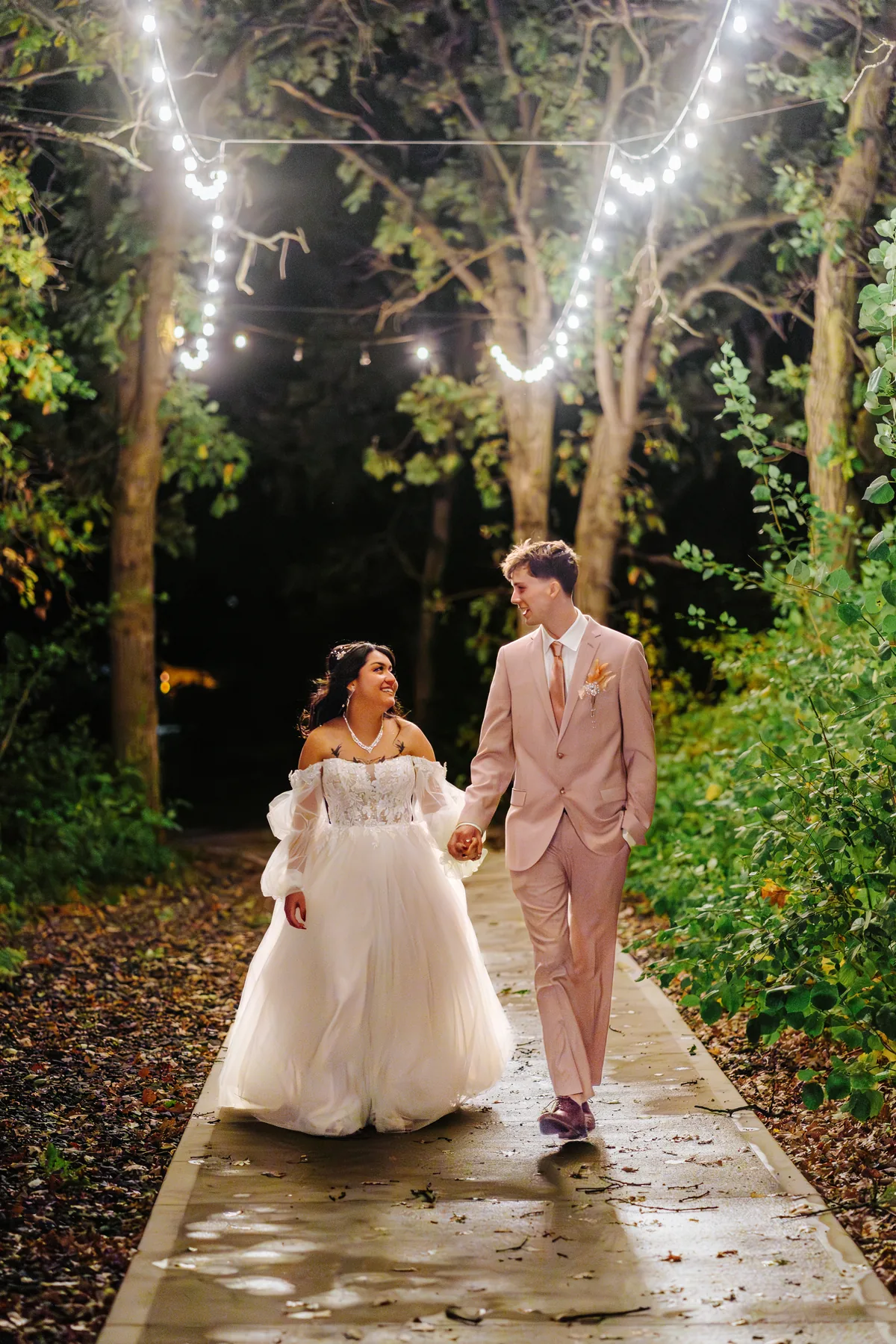 Paola and Riley walking the tree path under string lights at night at Oakland Estates Manitoba wedding — Chris Ngo