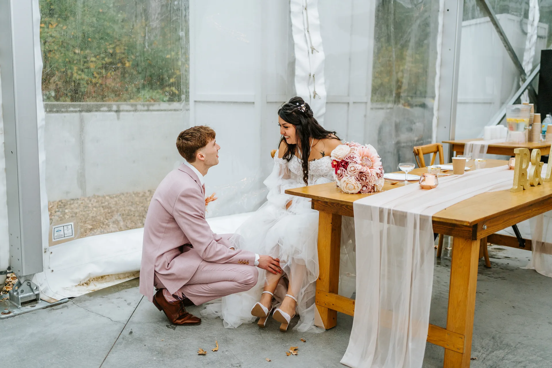 Groom Riley kneeling to put bride Paola's shoe on during reception — candid moment at Oakland Estates Manitoba wedding