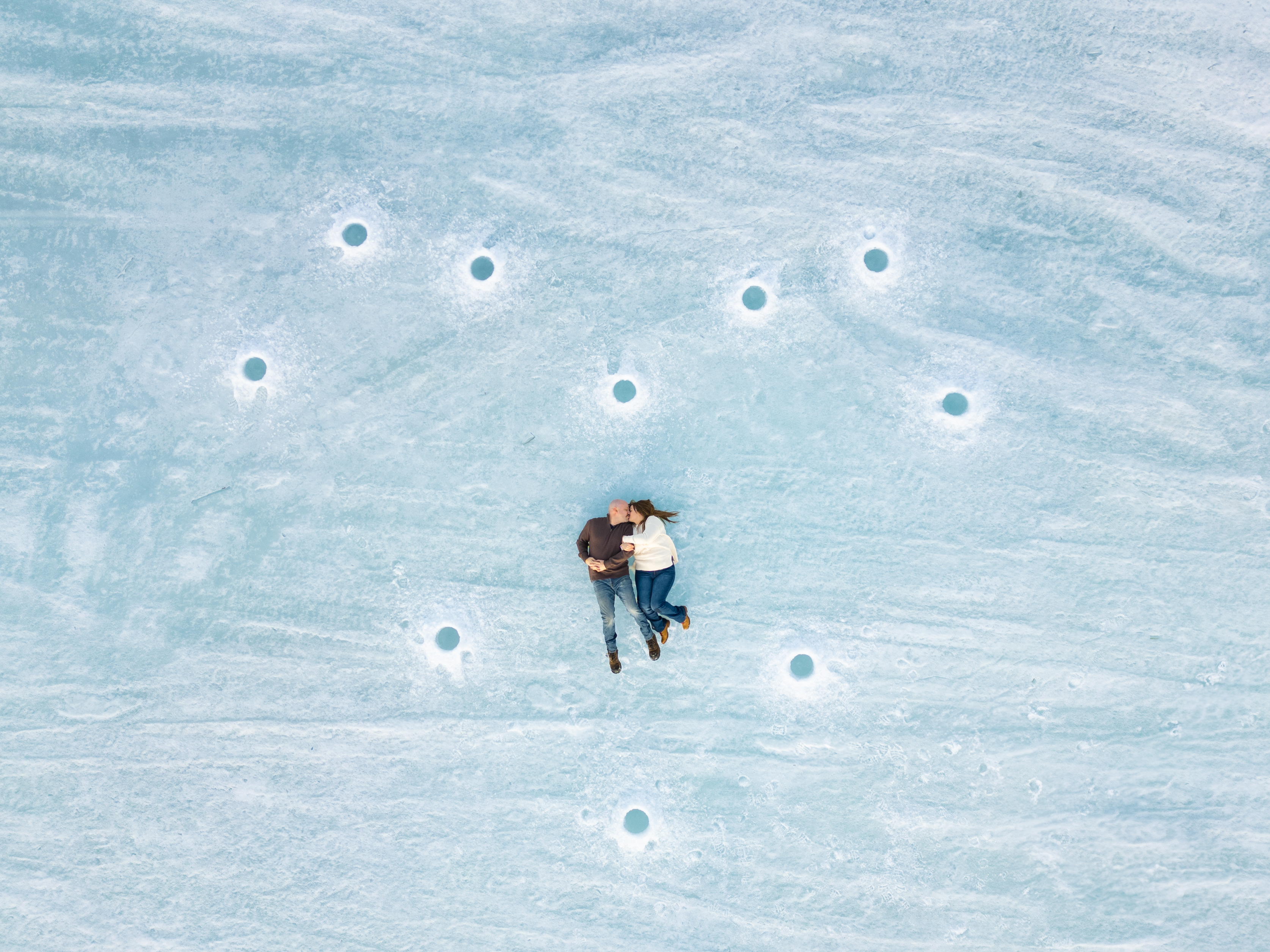 Aerial drone photo of Ryan and Vanessa lying together on a frozen Manitoba lake surrounded by ice fishing holes in the snow — winter engagement photography by Ngo Photography Winnipeg