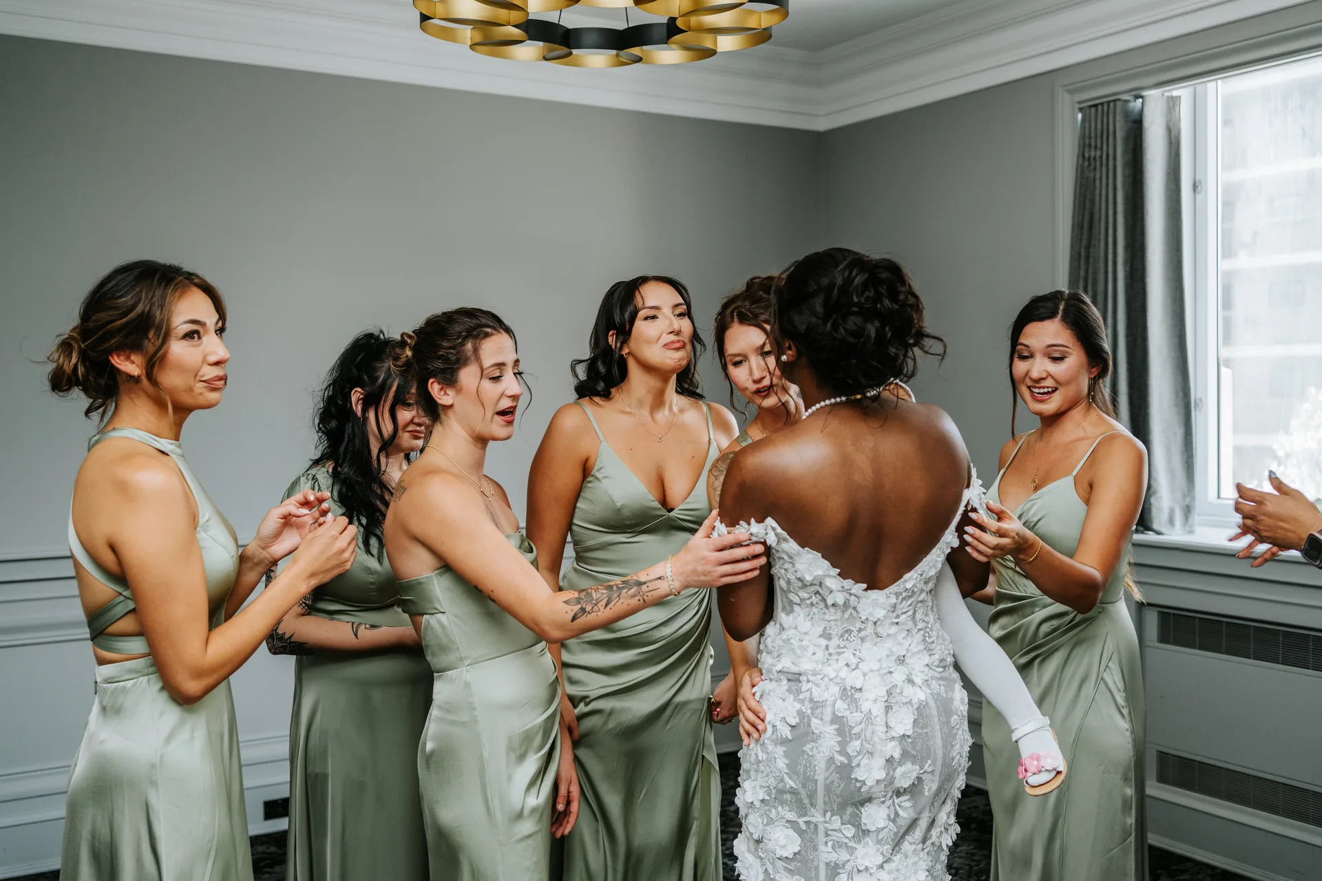Bridesmaids in sage green dresses helping button up the bride's gown at the Fort Garry Hotel, Winnipeg