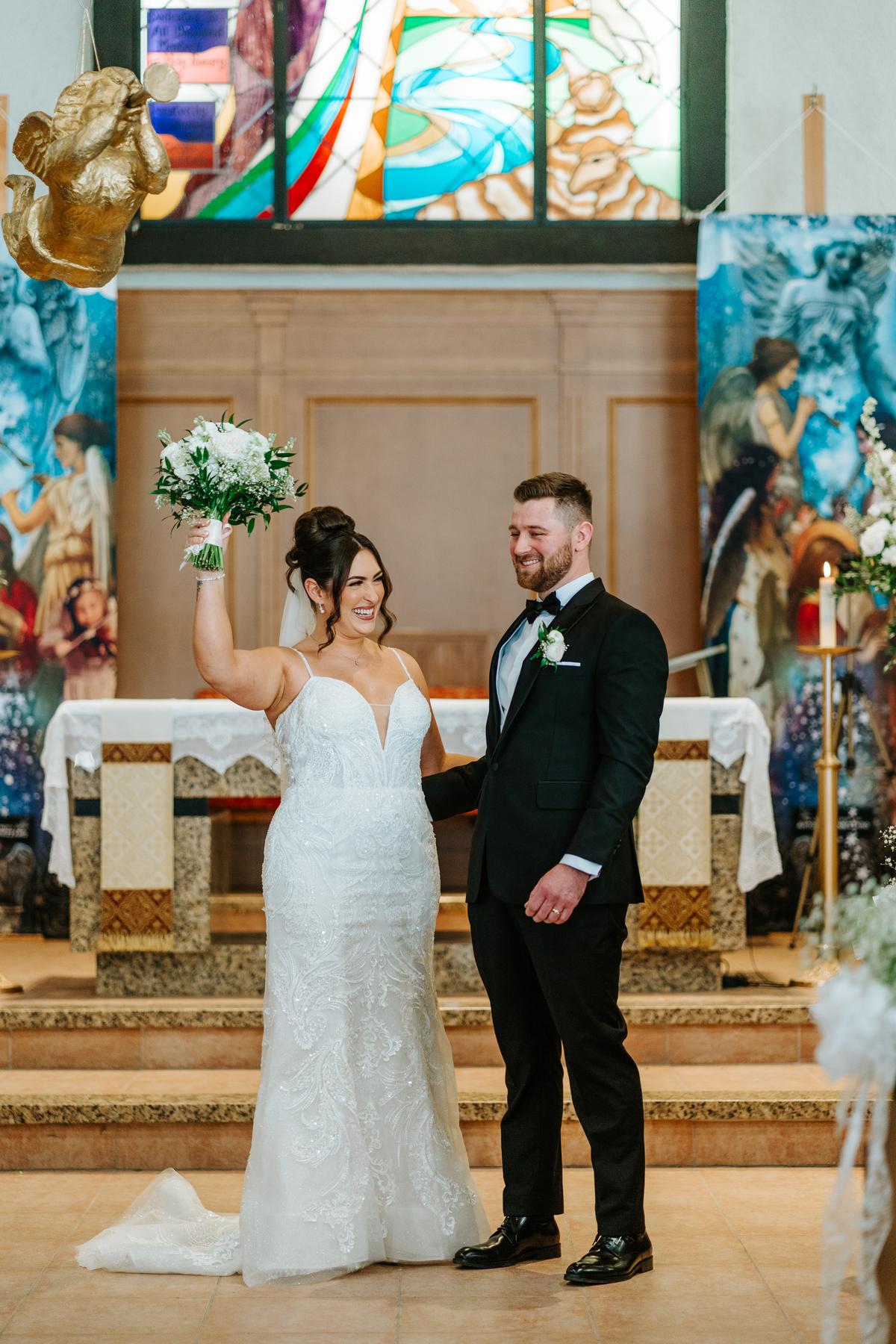 Stef raising her bouquet in celebration after the recessional at Holy Rosary Church Winnipeg, the congregation cheering — wedding photography by Ngo Photography