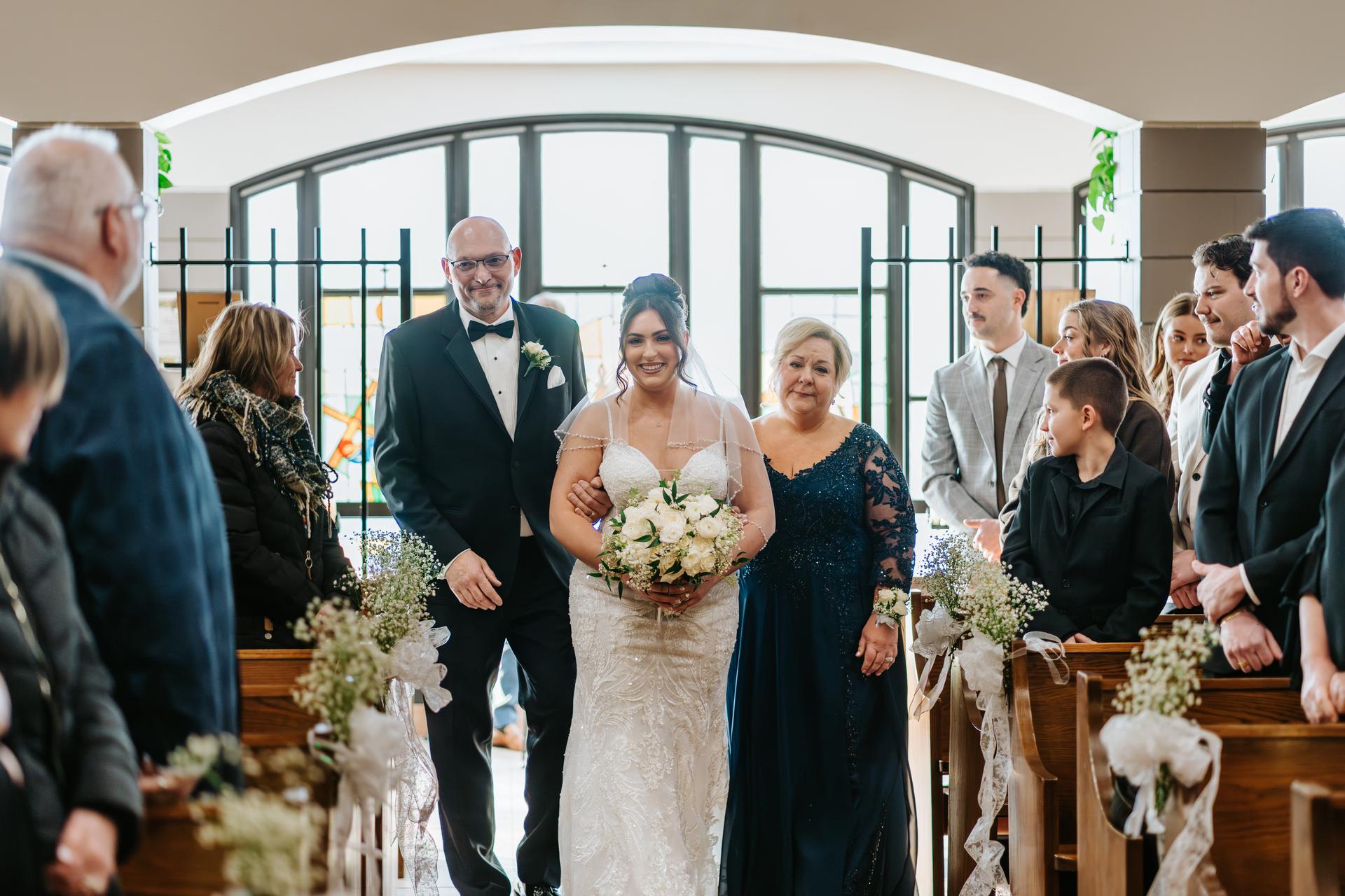 Stef walking down the aisle with her parents at Holy Rosary Church, the congregation standing on either side — bridal processional photography by Ngo Photography Winnipeg