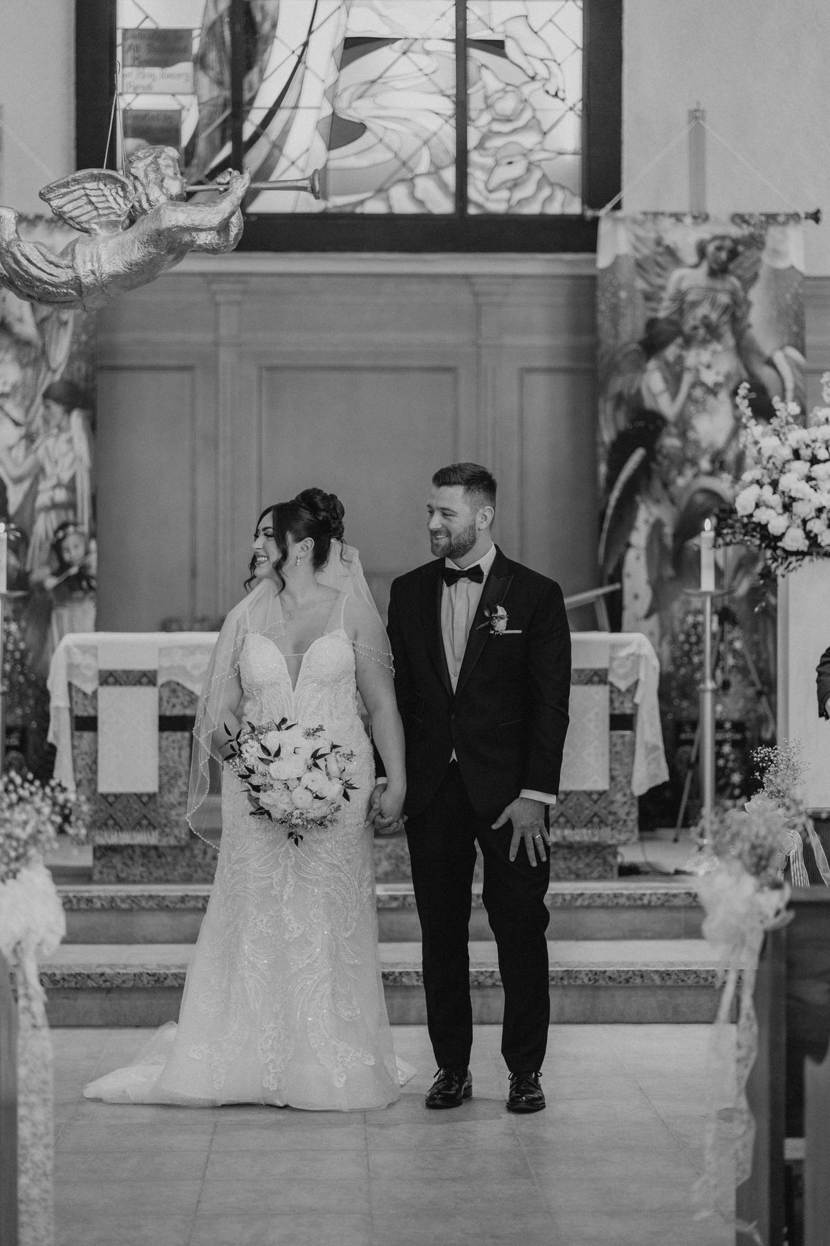 Stef and Brett standing together at the altar at Holy Rosary Church Winnipeg in black and white — wedding portrait by Ngo Photography