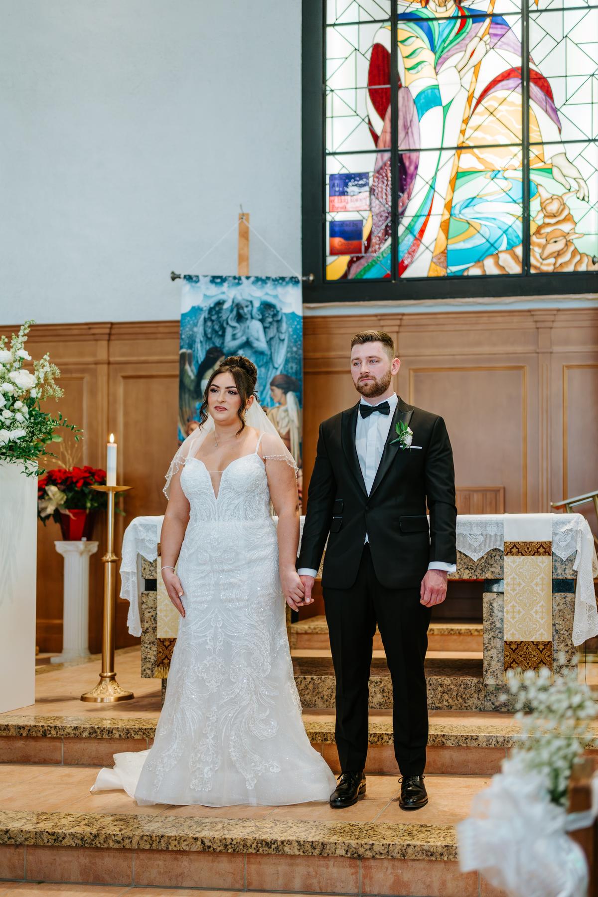 Stef and Brett holding hands at the altar at Holy Rosary Church, the colourful stained glass backdrop behind them — Catholic wedding photography Winnipeg
