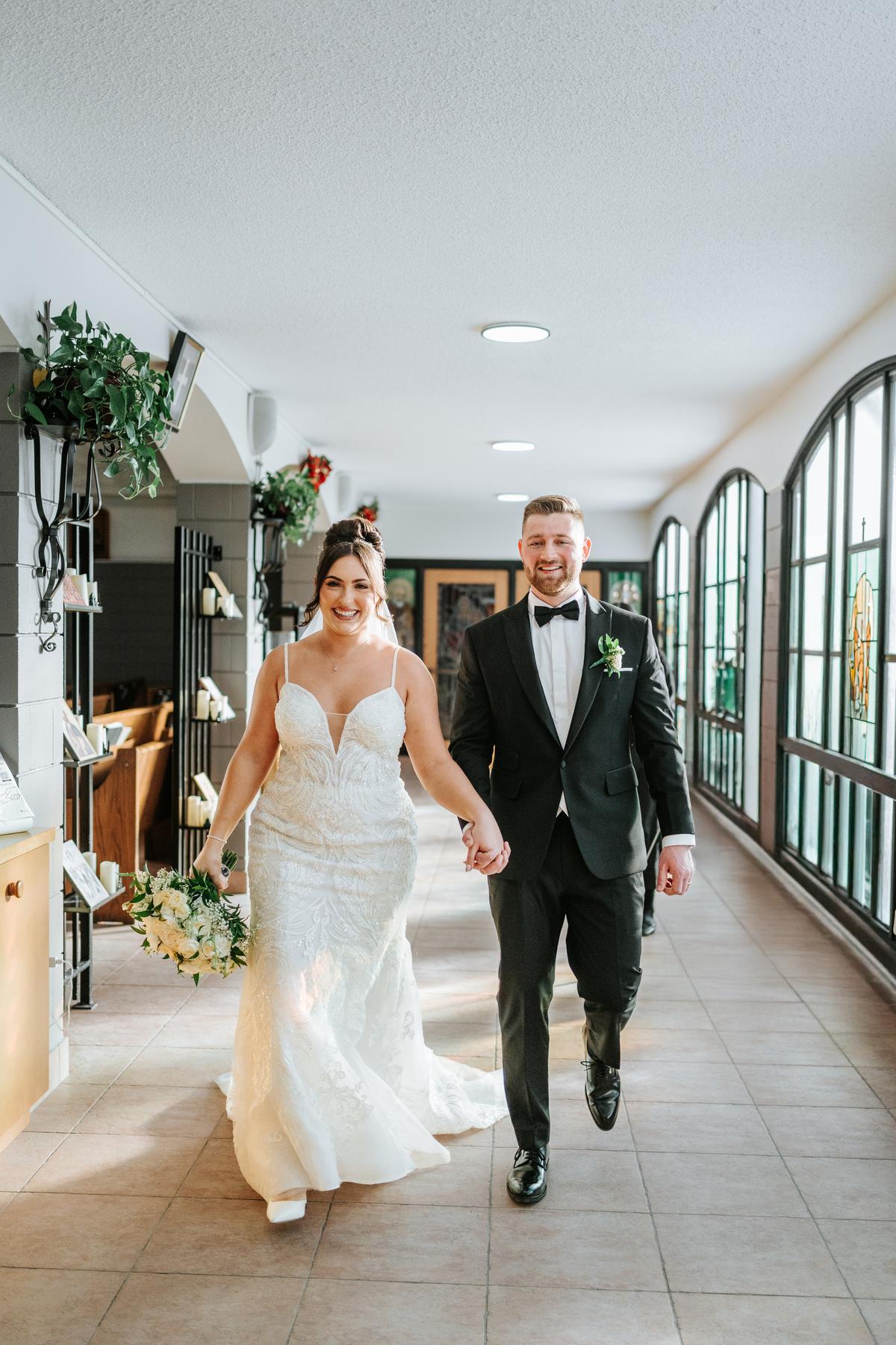 Stef and Brett walking through the bright narthex foyer at Holy Rosary Church after the ceremony, hand in hand — wedding photography by Ngo Photography Winnipeg