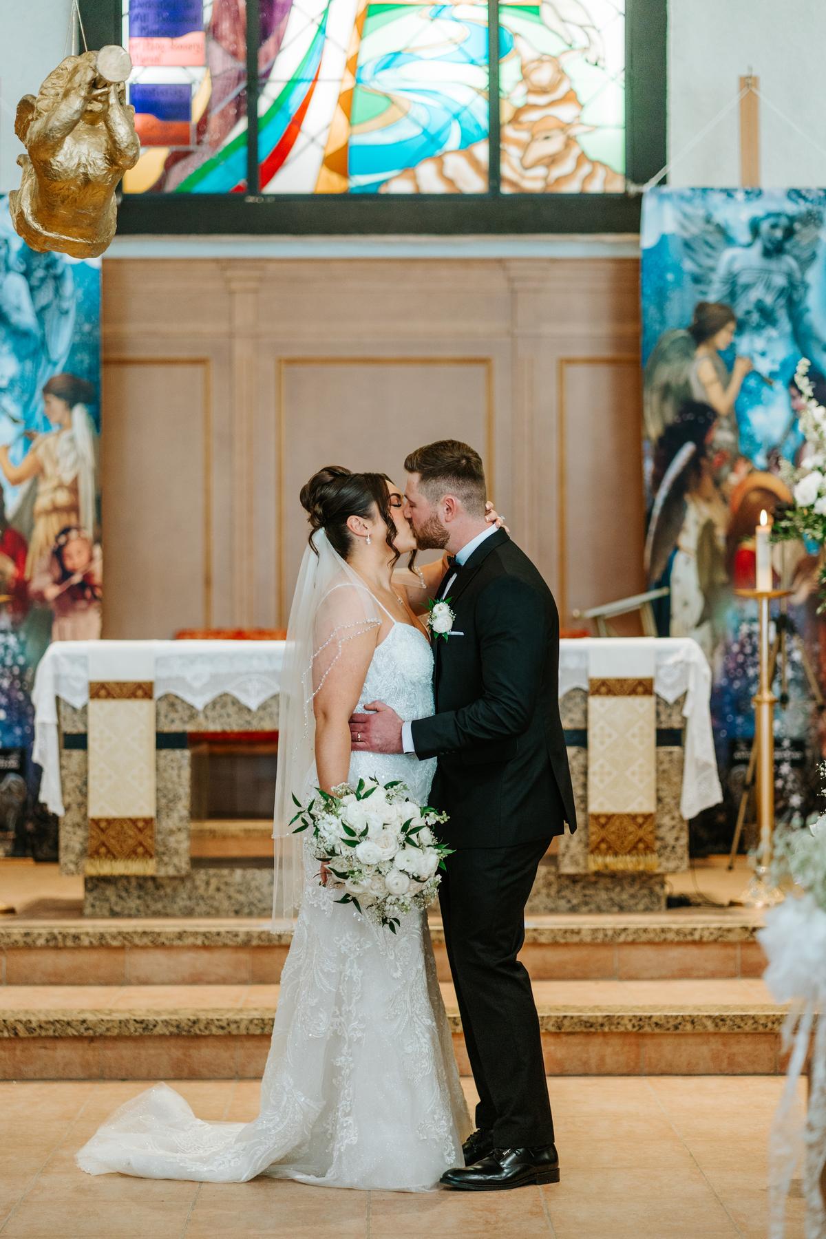 Stef and Brett's first kiss at the altar at Holy Rosary Church Winnipeg, the vibrant stained glass glowing behind them — wedding ceremony photography by Ngo Photography