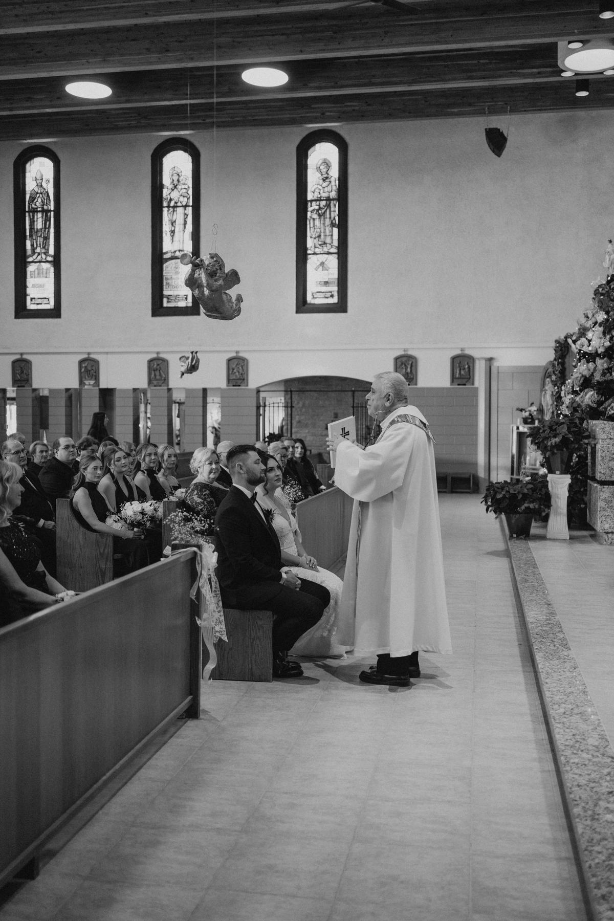 Brett kneeling to receive a blessing from the priest at the altar at Holy Rosary Church Winnipeg — Catholic wedding ceremony photography by Ngo Photography