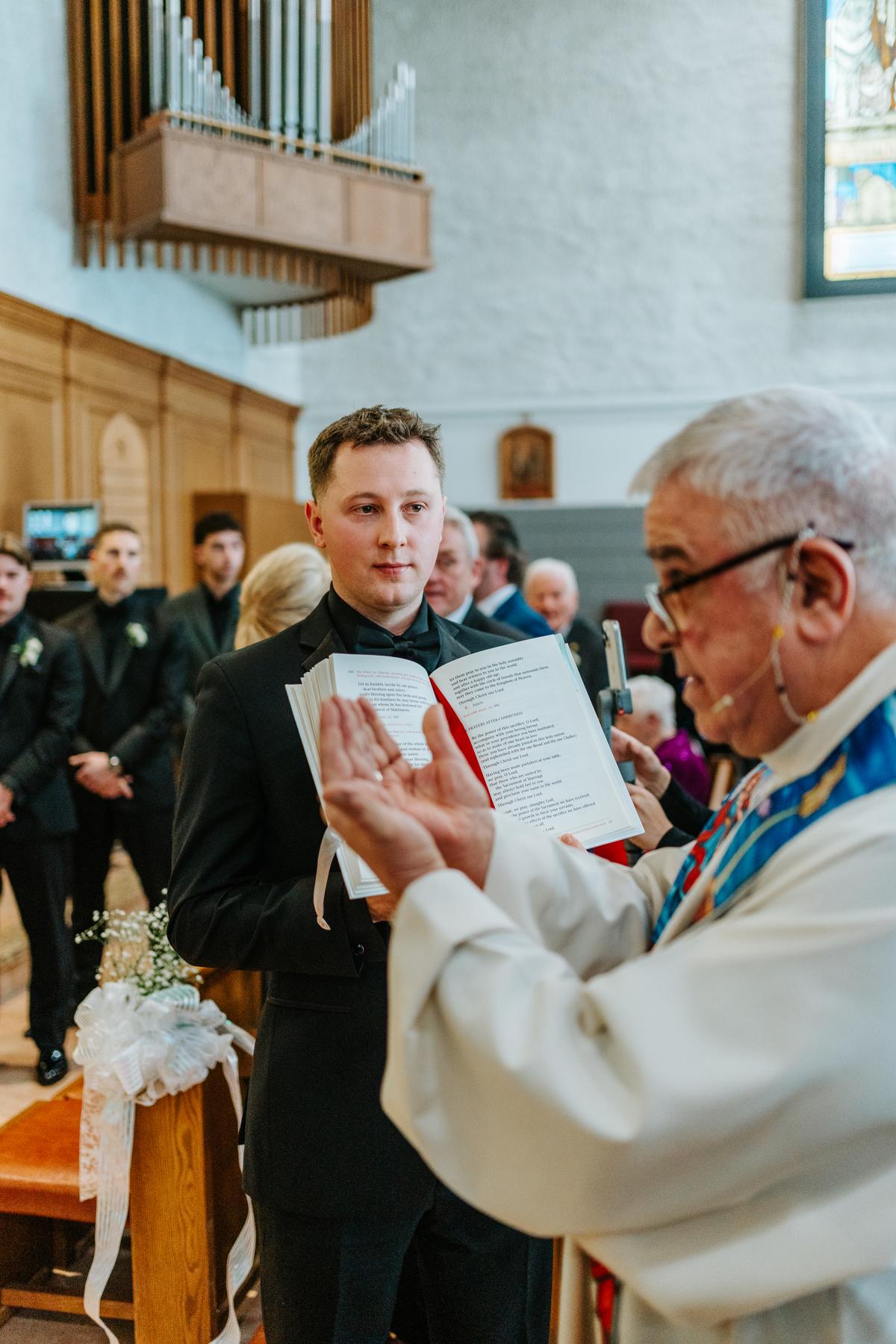 Brett receiving a blessing from the priest during the Catholic ceremony at Holy Rosary Church Winnipeg — wedding photography by Ngo Photography