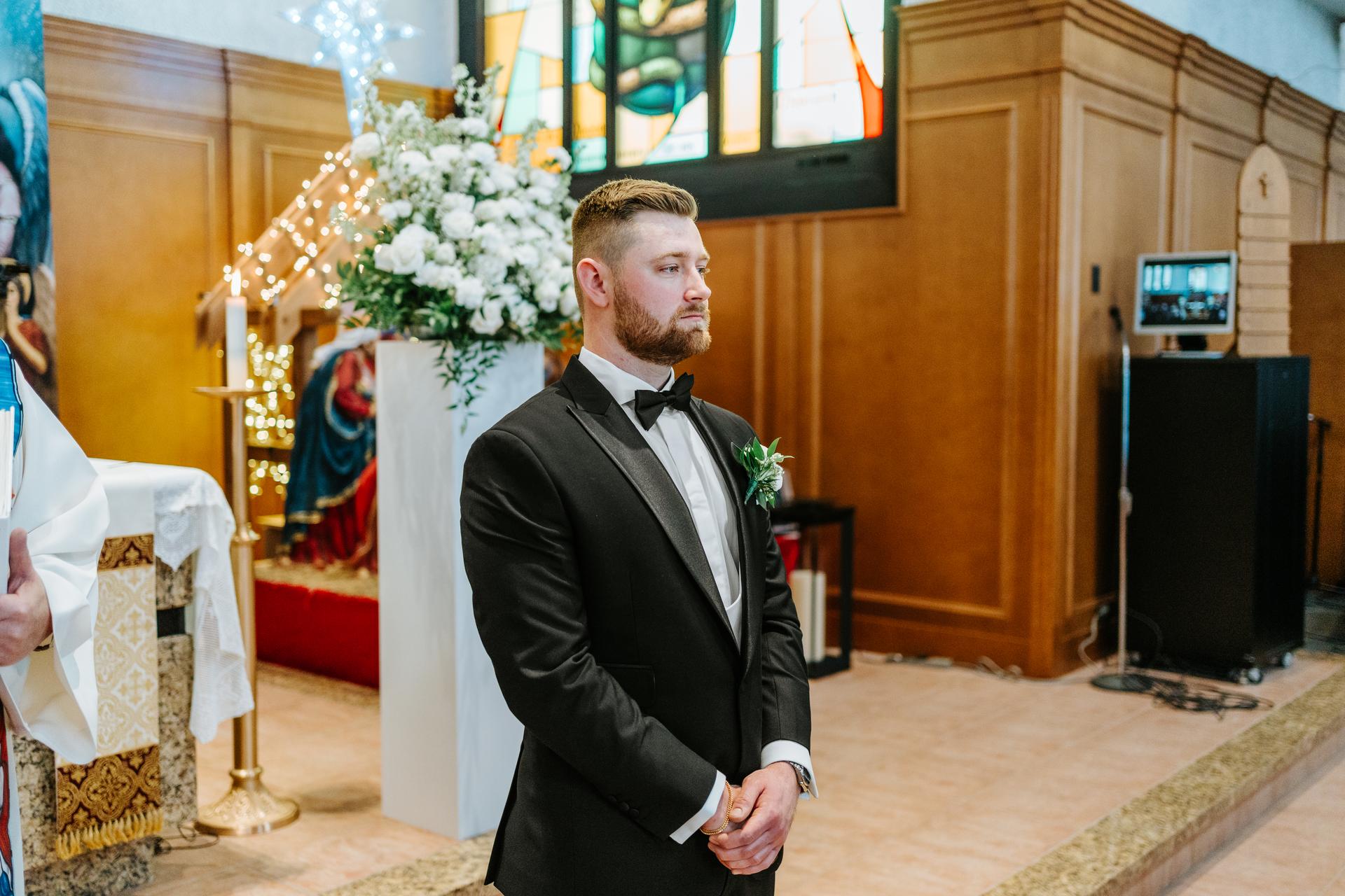 Brett waiting at the altar at Holy Rosary Church, the vibrant stained glass windows glowing behind him — wedding photography by Ngo Photography Winnipeg