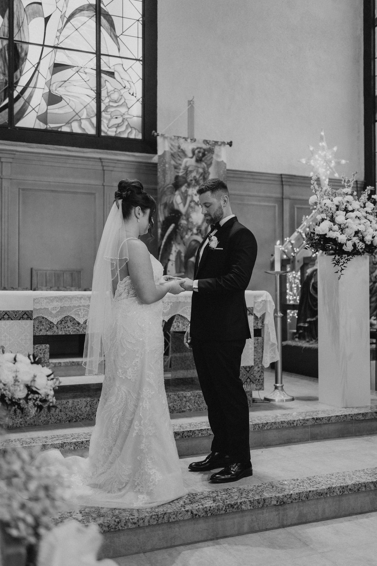 Stef placing Brett's ring on his finger during the ring exchange at Holy Rosary Church Winnipeg — black and white wedding ceremony photography by Ngo Photography