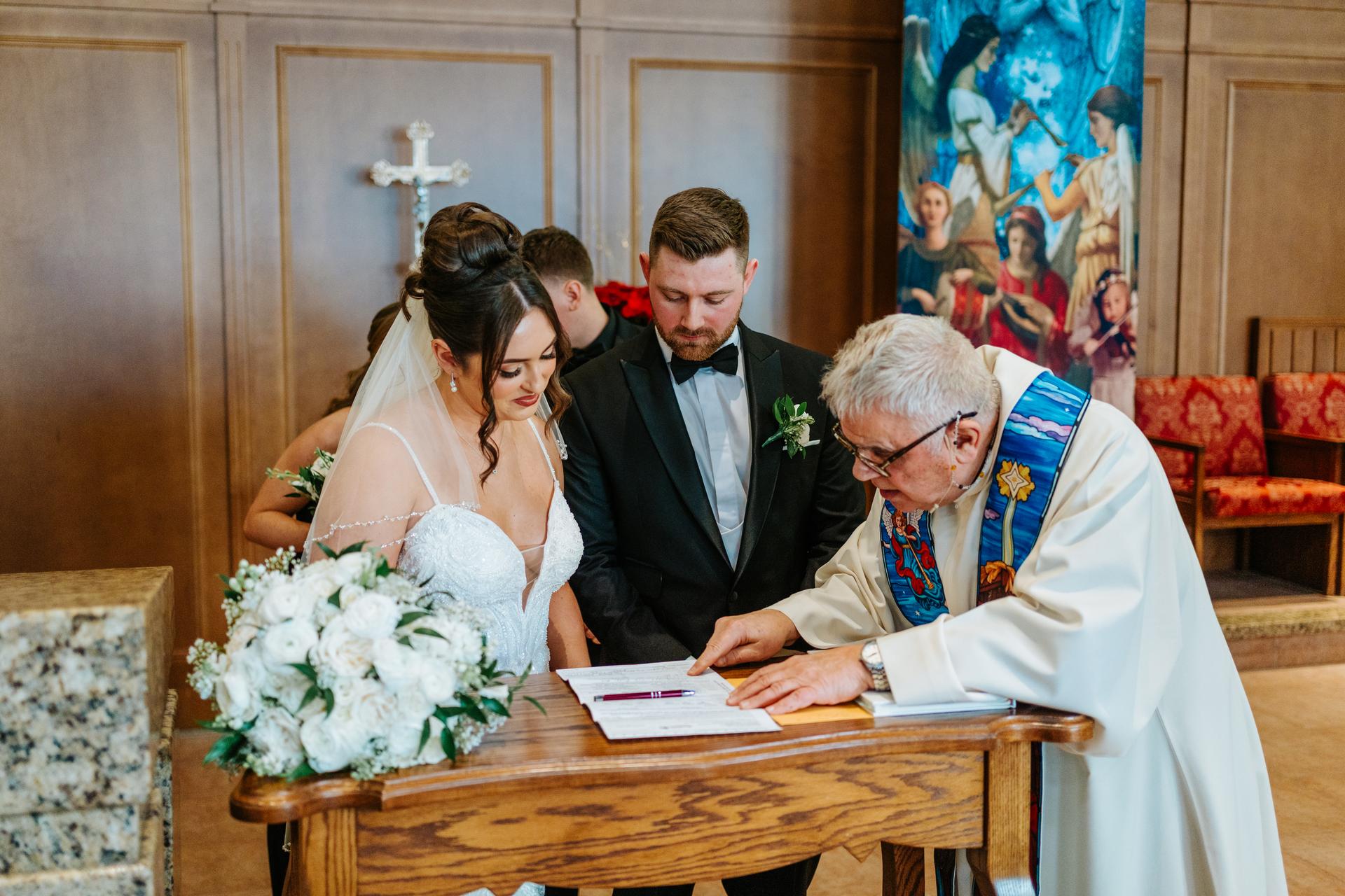 Stef signing the marriage register with Brett and the priest at Holy Rosary Church Winnipeg — Catholic wedding ceremony documentation by Ngo Photography