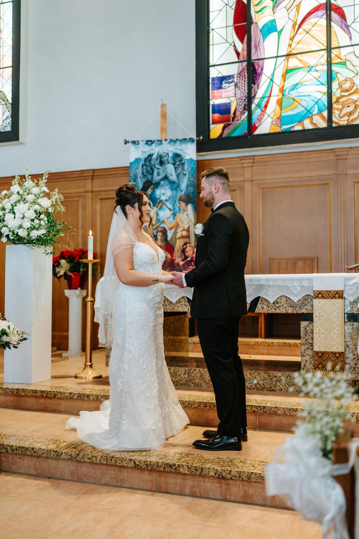 Stef and Brett exchanging vows at the altar at Holy Rosary Church, stained glass glowing behind them — wedding photography by Ngo Photography Winnipeg