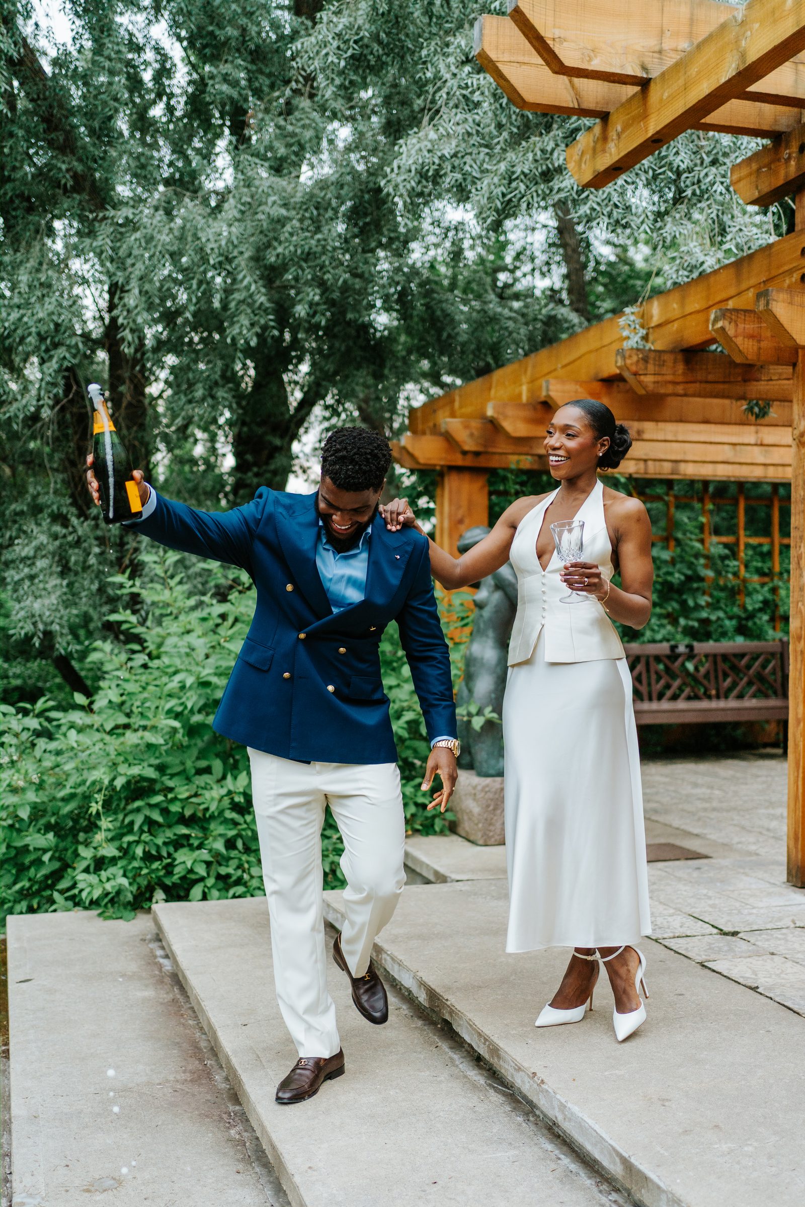 Emeka popping a champagne bottle outside the wooden pergola at Leo Mol Sculpture Garden in Assiniboine Park, Winnipeg — Tobi beside him holding a crystal flute, both laughing — post-ceremony celebration photo by Ngo Photography