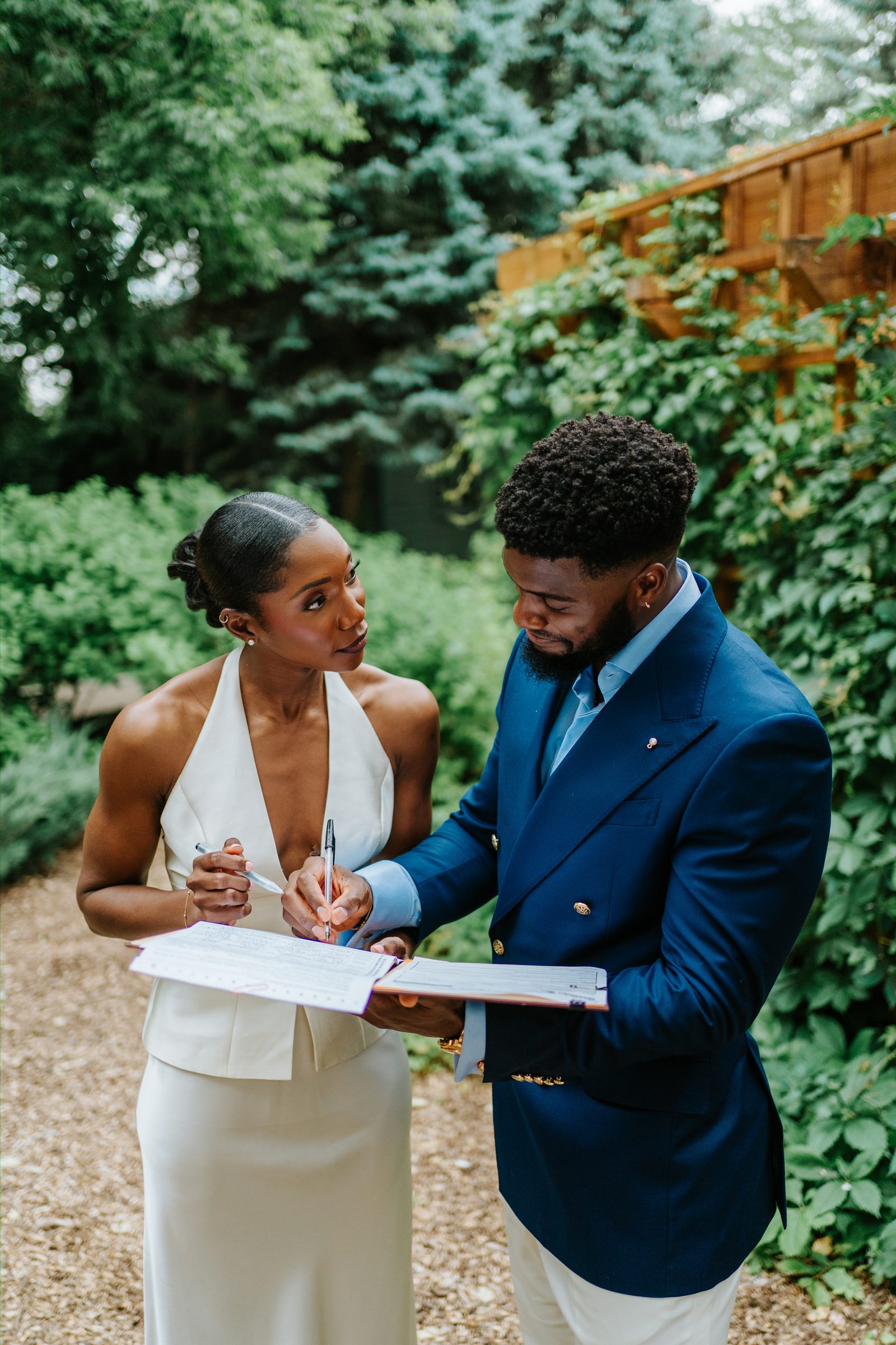 Tobi and Emeka signing the marriage licence together at Leo Mol Sculpture Garden in Assiniboine Park, Winnipeg — Tobi looks at Emeka as he signs, ivy and garden greenery in the background — documentary wedding photography by Chris Ngo, Ngo Photography