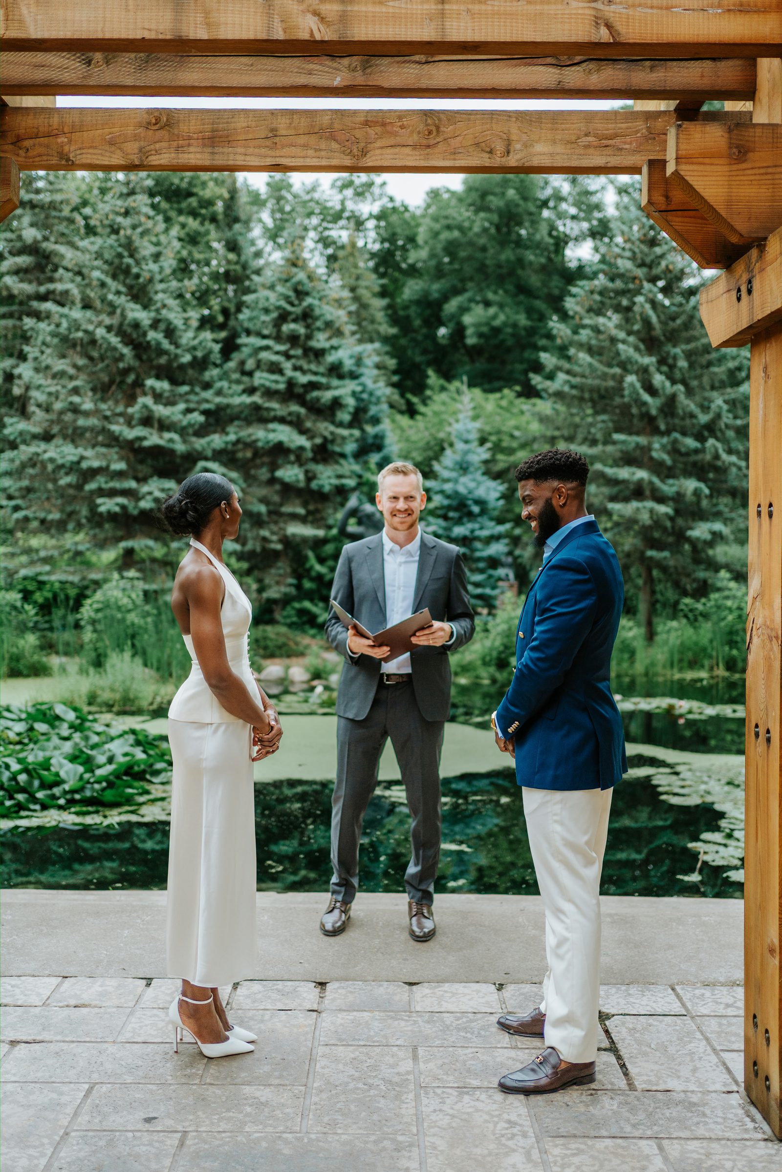 Tobi and Emeka exchanging vows under the wooden pergola at Leo Mol Sculpture Garden in Assiniboine Park, Winnipeg — the officiant stands between them, the lily pond with lily pads visible behind — intimate micro-wedding ceremony photographed by Chris Ngo, Ngo Photography