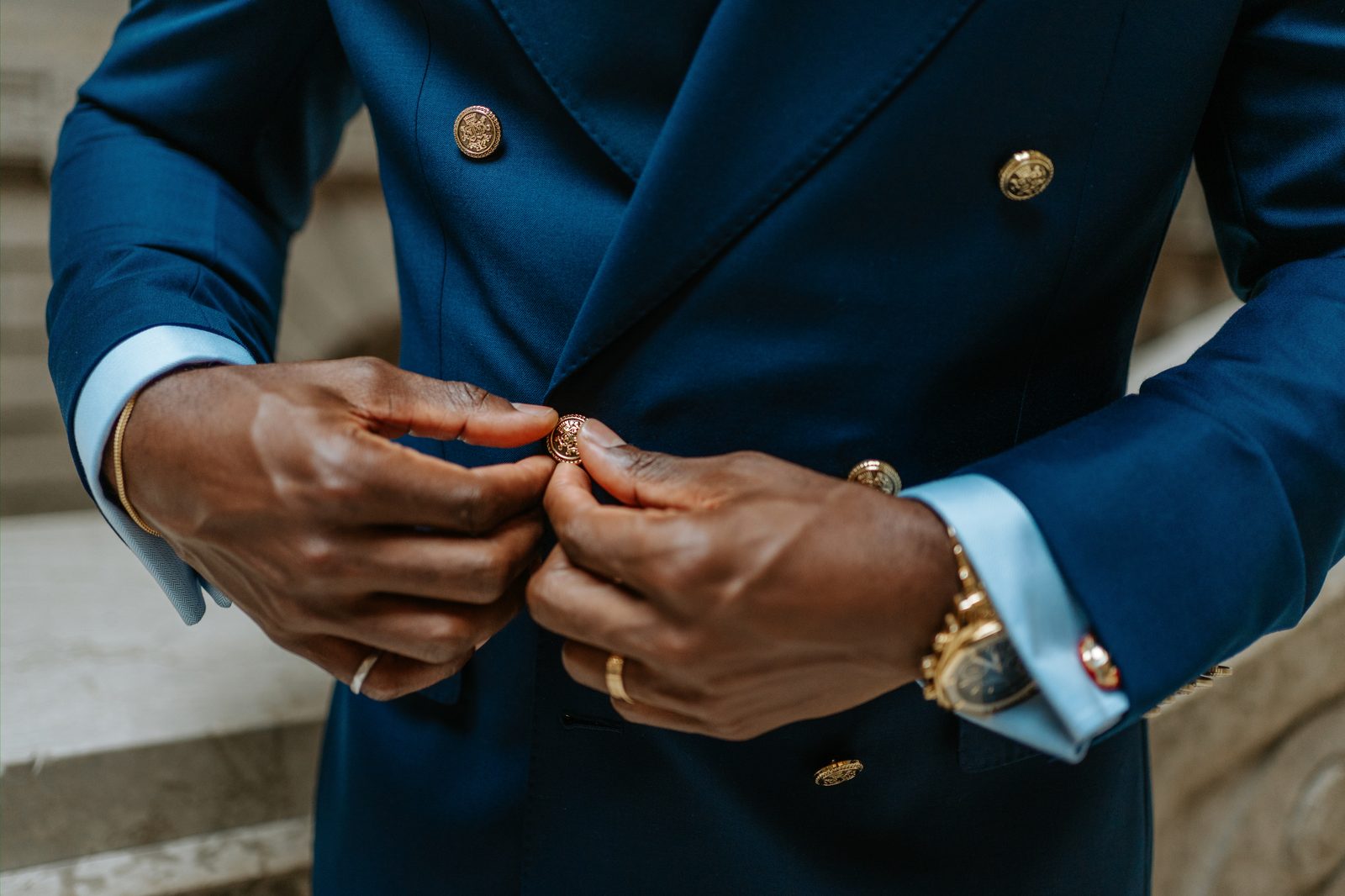 Close detail of Emeka's hands buttoning his navy double-breasted suit jacket — gold cufflinks, gold Rolex watch, gold ring, light blue shirt cuffs — on the Legislative Building marble steps, Winnipeg
