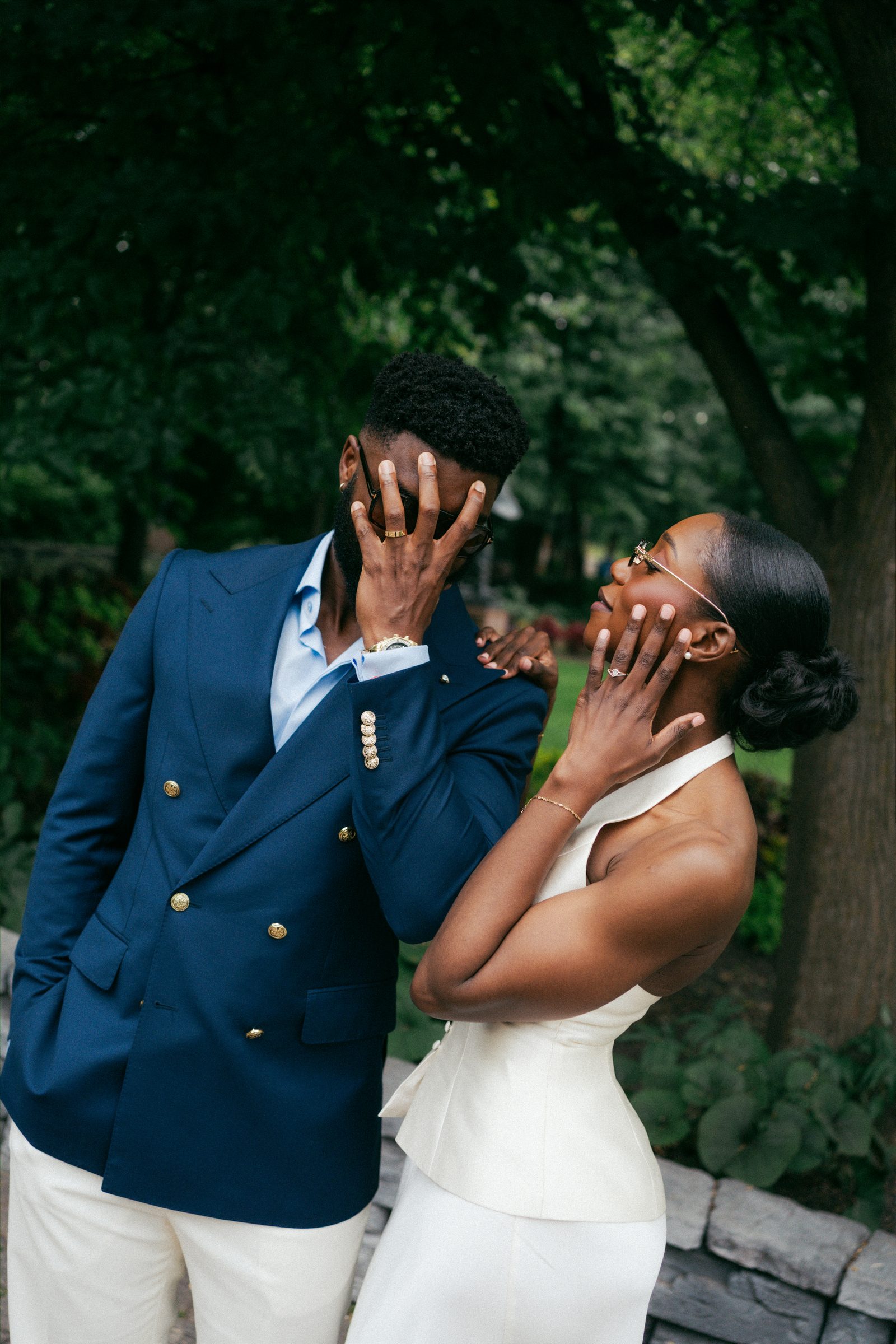 Film-style black and white portrait of Tobi and Emeka covering their mouths with their hands in an emotional, overwhelmed reaction — both laughing — at Leo Mol Sculpture Garden, Assiniboine Park, Winnipeg, by Ngo Photography