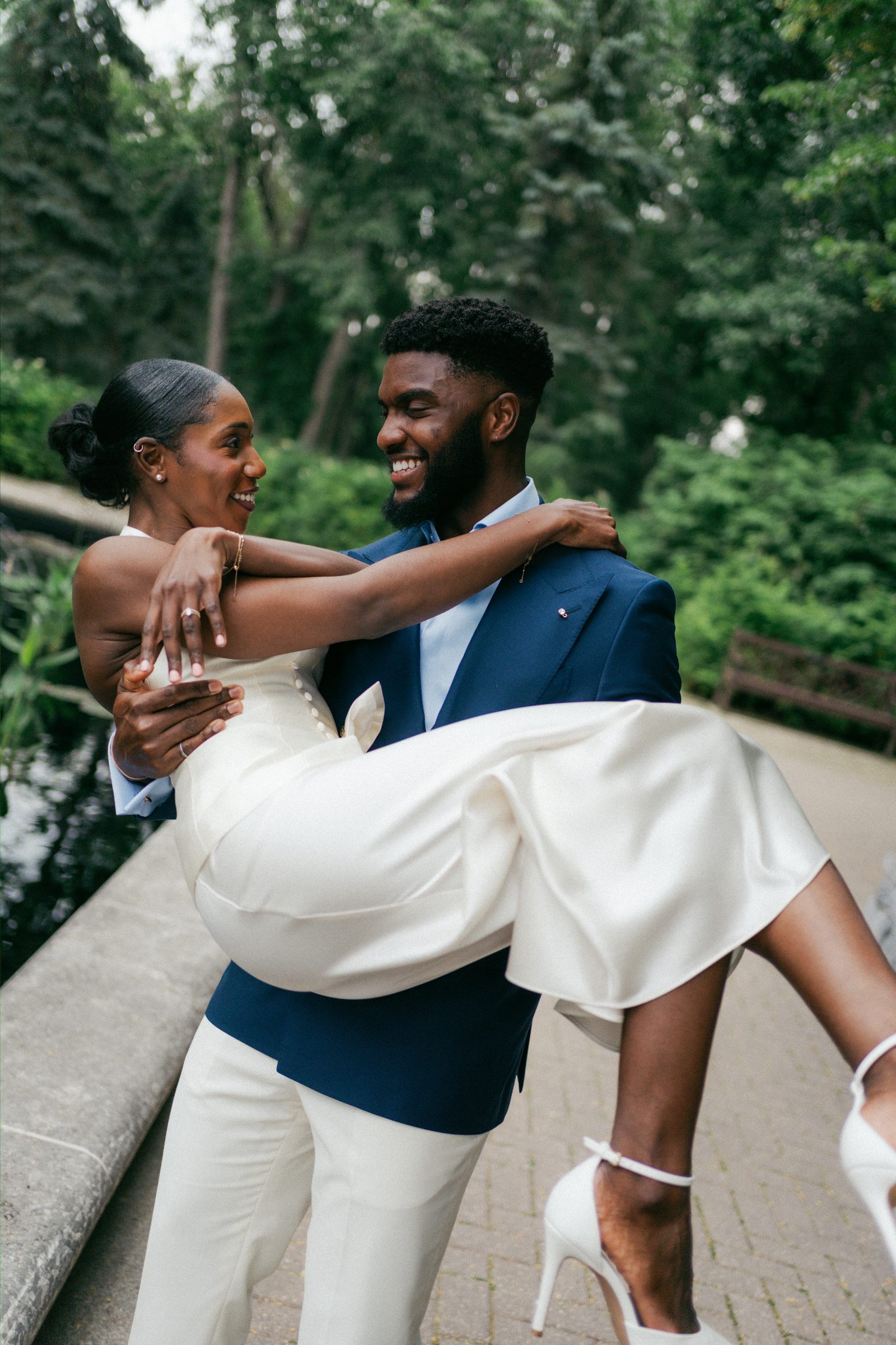 Film-style colour portrait of Tobi and Emeka both covering their faces with their hands in a laughing, overwhelmed moment — wedding rings visible — at Leo Mol Sculpture Garden in Assiniboine Park, Winnipeg — post-ceremony portrait by Chris Ngo, Ngo Photography