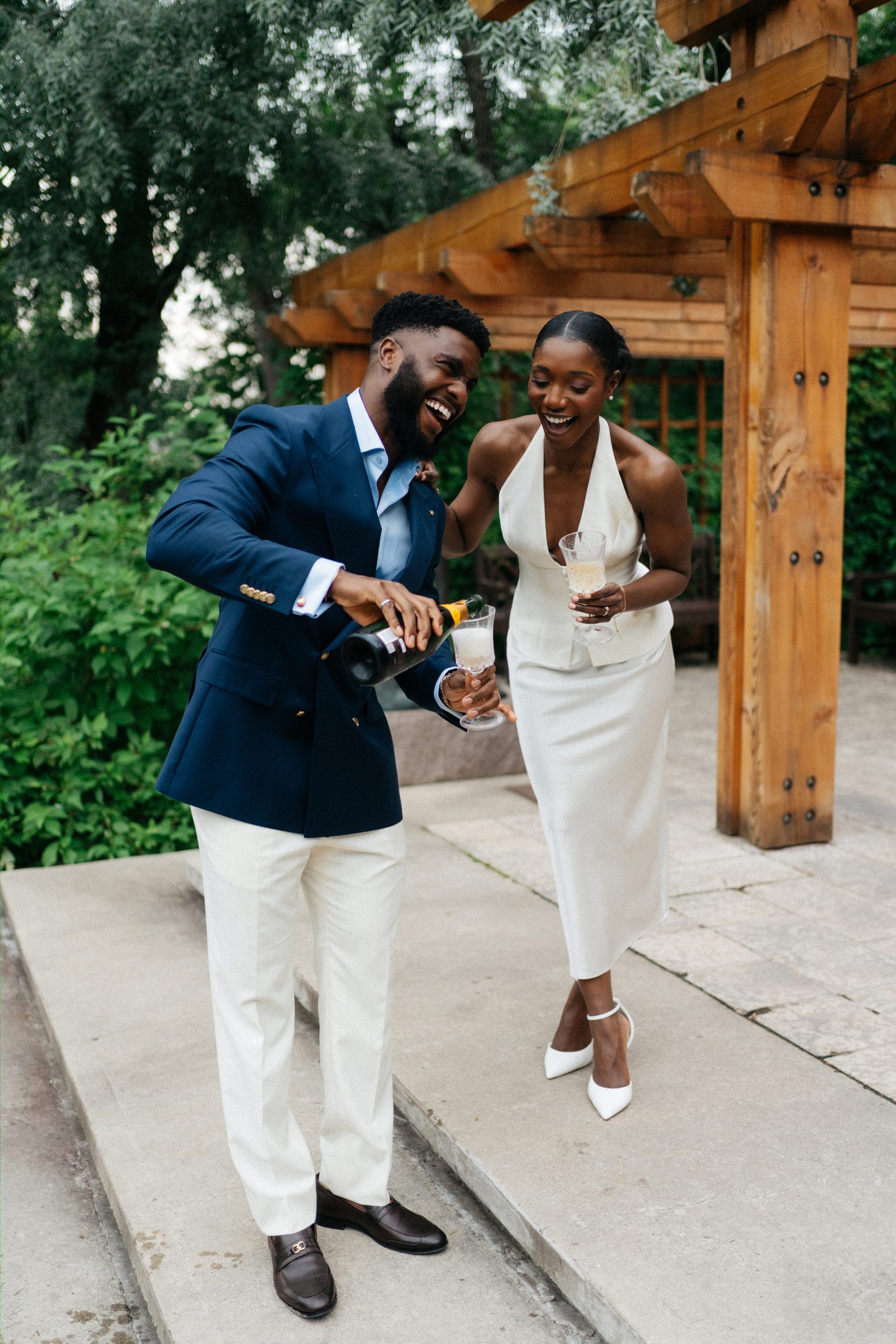 Film-style portrait of Emeka pouring champagne for Tobi at Leo Mol Sculpture Garden — both laughing, the wooden pergola behind them — Assiniboine Park, Winnipeg
