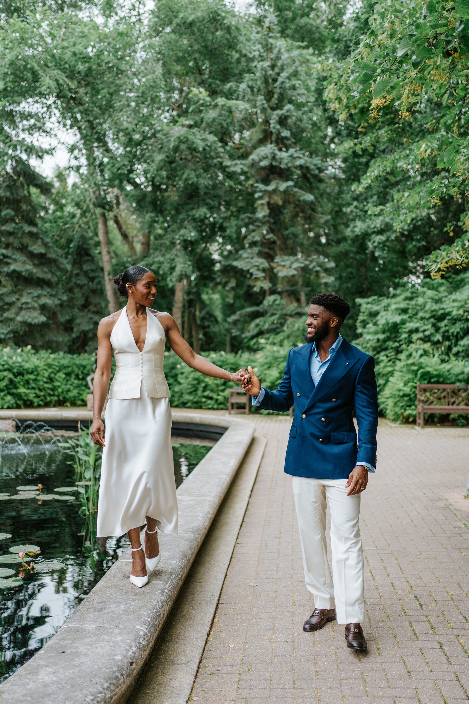 Tobi walking along the edge of the lily pond at Leo Mol Sculpture Garden in Assiniboine Park, Winnipeg — Emeka holding her hand from the garden path — post-ceremony portrait by Ngo Photography