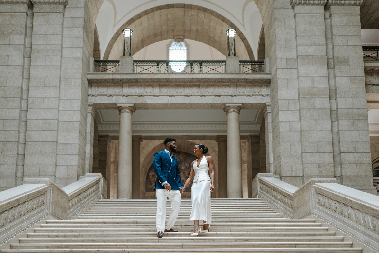 Tobi and Emeka holding hands on the grand marble staircase inside the Manitoba Legislative Building, Winnipeg — Emeka in a navy double-breasted suit with gold buttons, Tobi in a white halter-neck two-piece with a blush bouquet, photographed by Ngo Photography