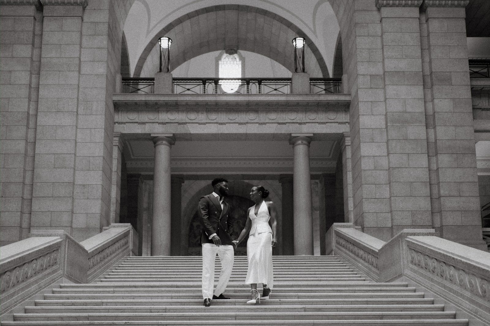 Tobi and Emeka walking down the grand staircase of the Manitoba Legislative Building — wide view showing the full sweep of marble and arched architecture behind them — Winnipeg wedding photographer Chris Ngo