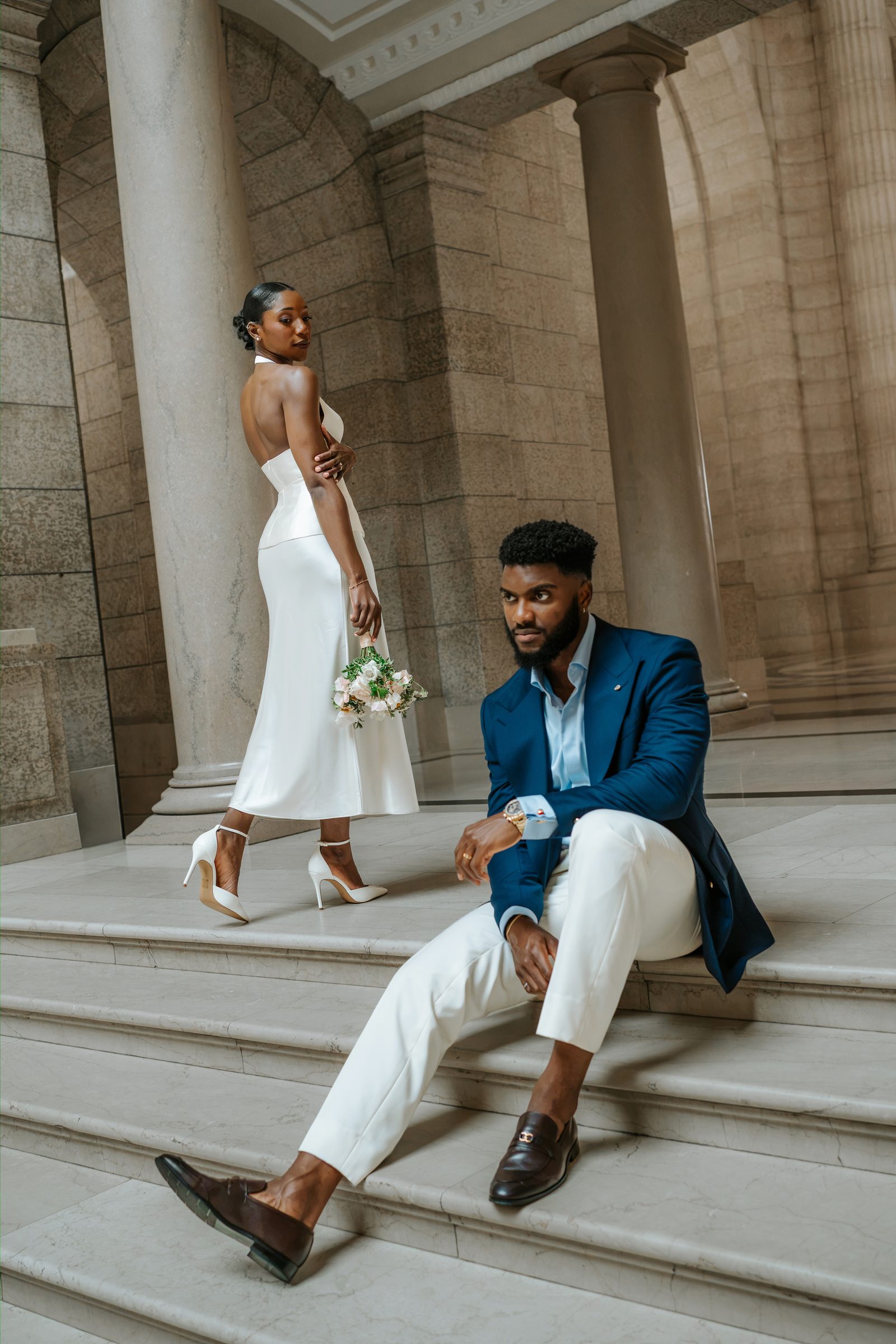 Emeka seated on the Legislative Building exterior marble steps, Tobi standing and looking over her shoulder with blush bouquet — editorial couple portrait between stone columns, Winnipeg wedding photographer Chris Ngo
