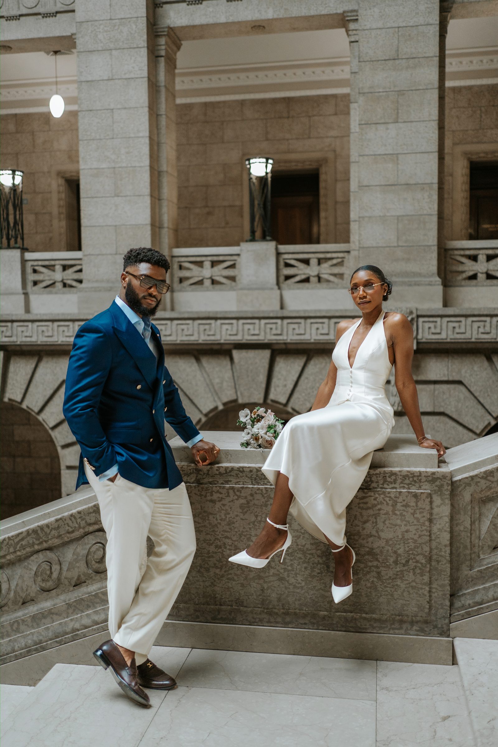 Tobi and Emeka inside the Manitoba Legislative Building rotunda — both wearing sunglasses, leaning against an ornate carved marble balustrade with Greek key frieze in the background — editorial couple portrait, Winnipeg wedding photographer Ngo Photography